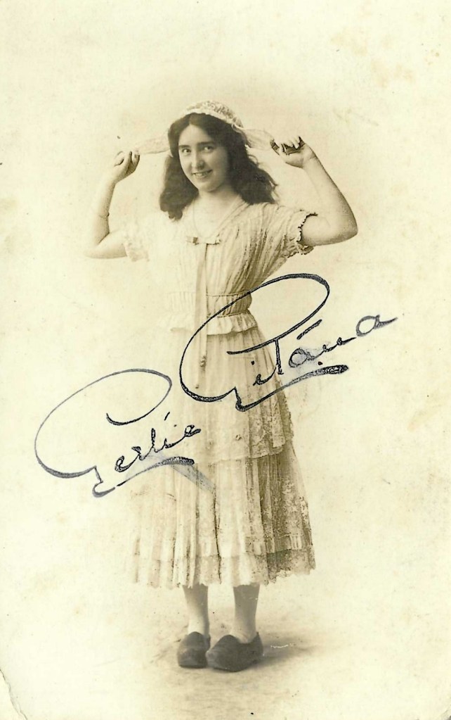 A vintage black and white photograph of a young girl with dark hair, wearing a light-colored dress and holding a hat or scarf above her head, smiling at the camera. Her name, 'Gertie Gitana,' is beautifully signed below her image.